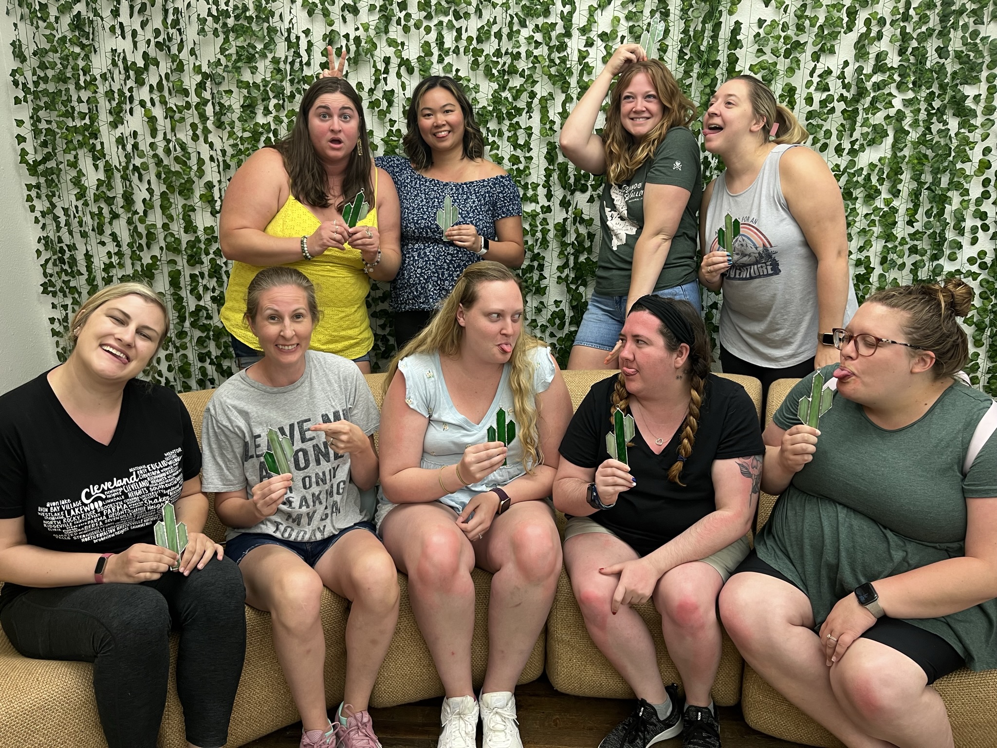 9 people smiling and making funny faces holding their stained glass cactus after a class at Foundry