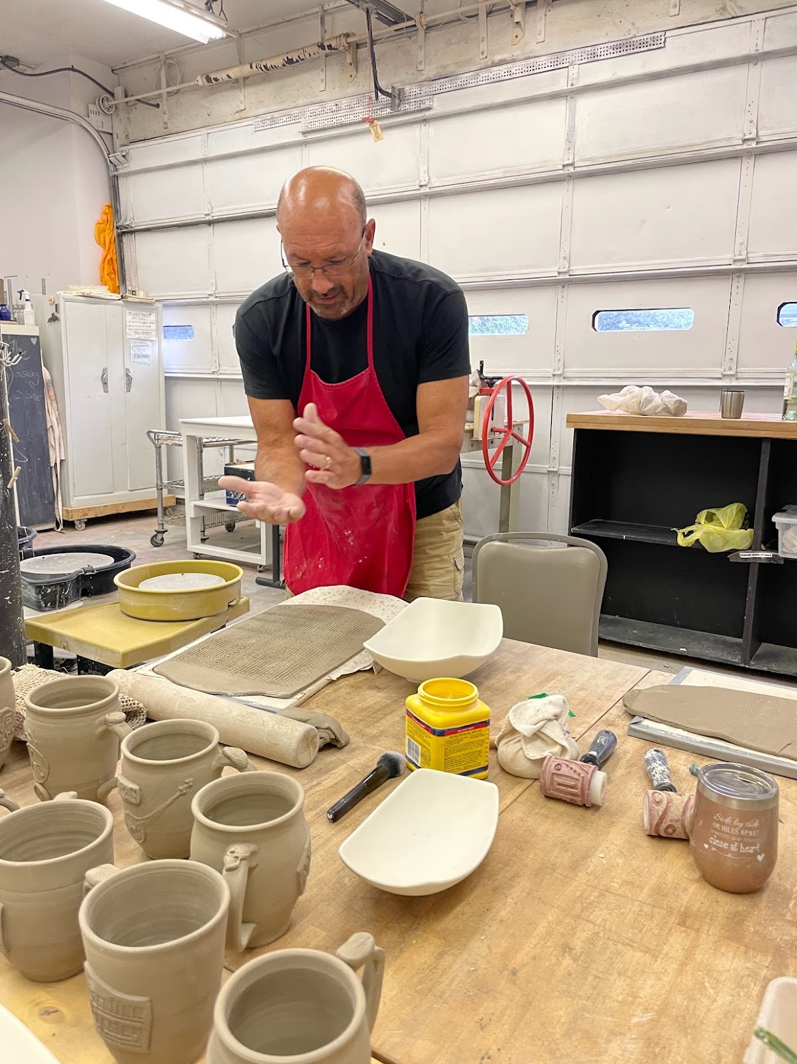 bald guy in red apron flattening clay to make a mug